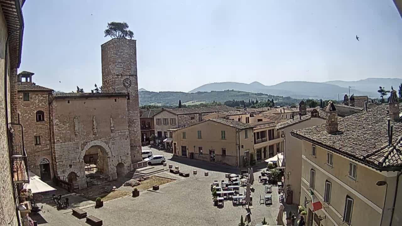 A town square in a hilly, sunny region, with stone buildings surrounding the plaza, one featuring a tall tower with a tree on top, and outdoor seating visible.