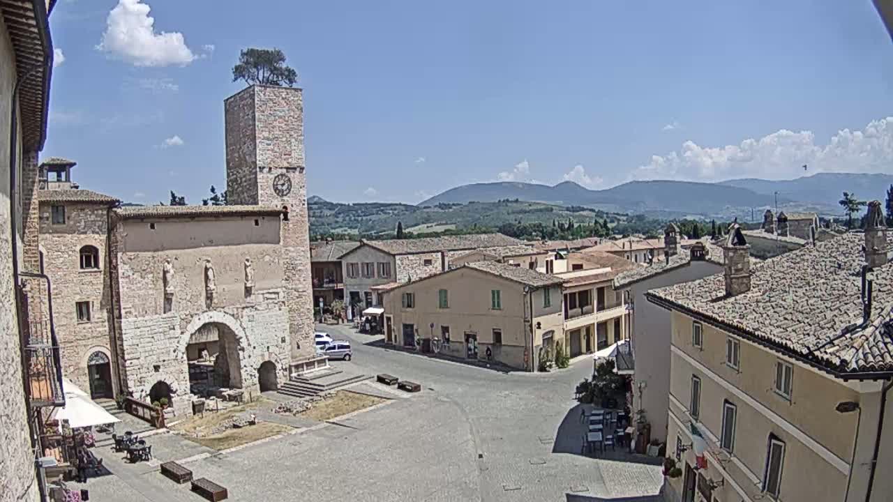 A town square in a small Italian town on a sunny day, with a tall stone clock tower, several buildings, and a view of rolling hills in the distance.