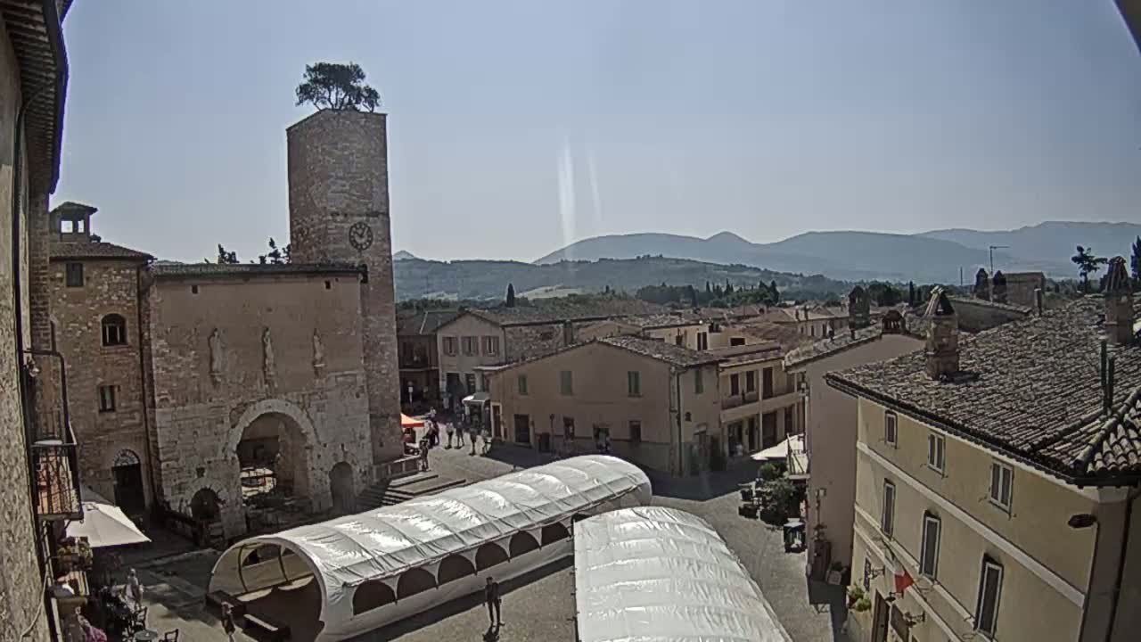 A sunny view of a town square in a hilltop town, featuring stone buildings, a clock tower with a tree on top, and several temporary canopies.