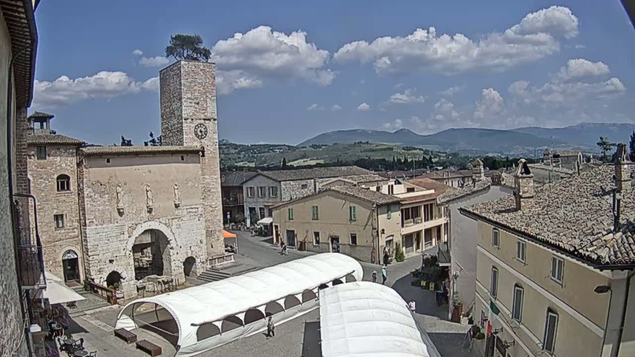 A town square with a stone tower and several buildings, under a partly cloudy sky, features white canopies in the square and distant hills visible in the background.