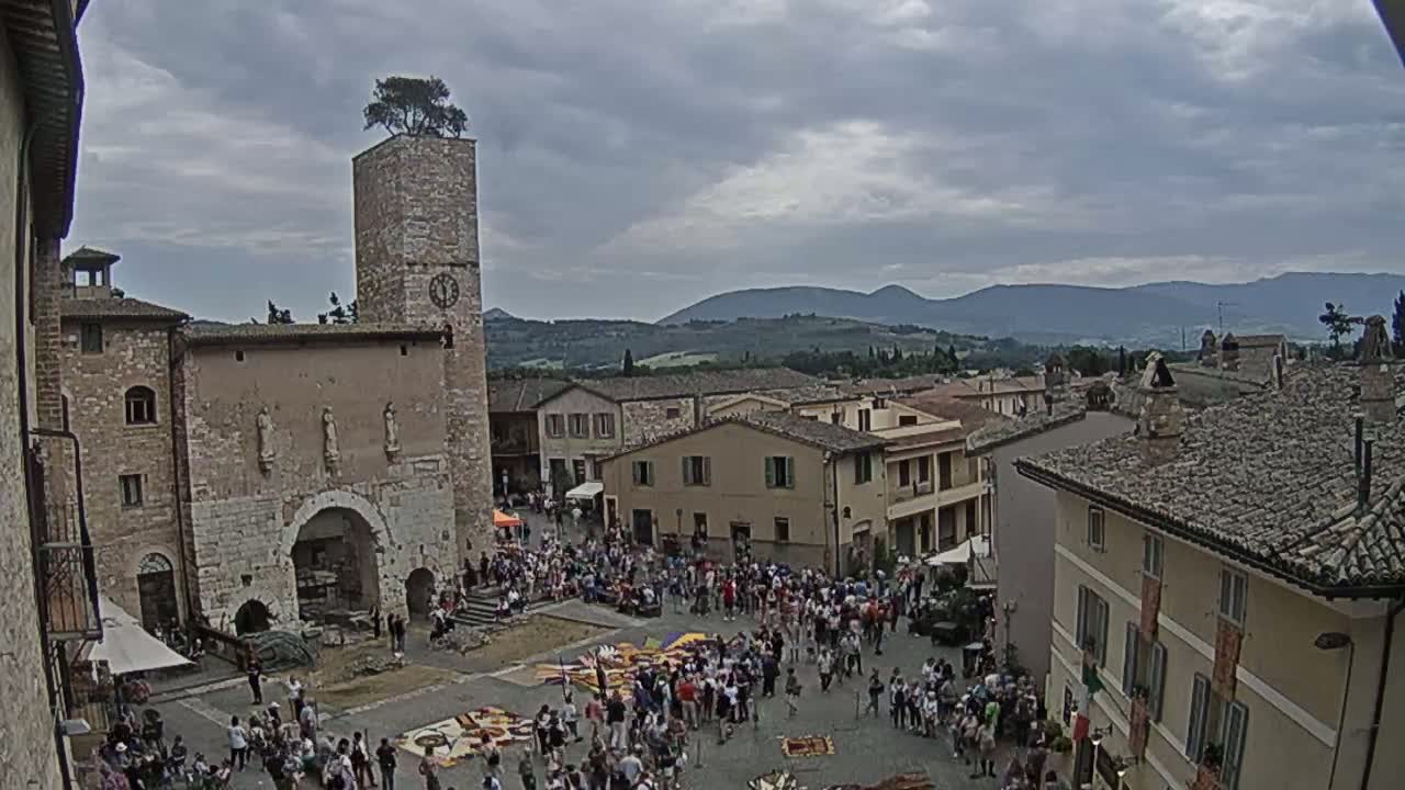 A town square filled with people under a cloudy sky, overlooked by buildings and a tall tower with a tree on top, with rolling hills visible in the distance.