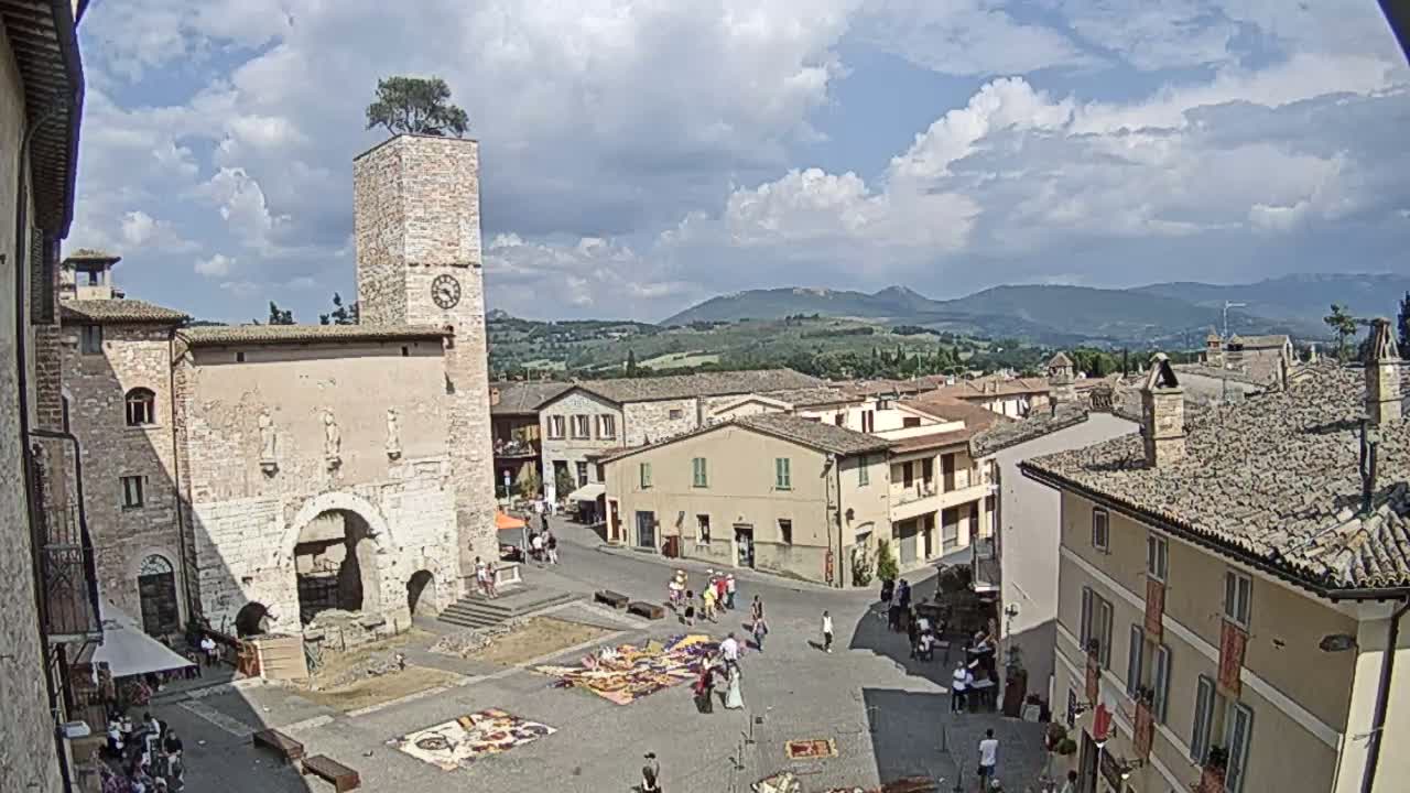 A town square, under a partly cloudy sky, is surrounded by stone buildings, one with a clock tower topped with a tree, and features people gathered in the square with colorful tapestries laid out on the ground.