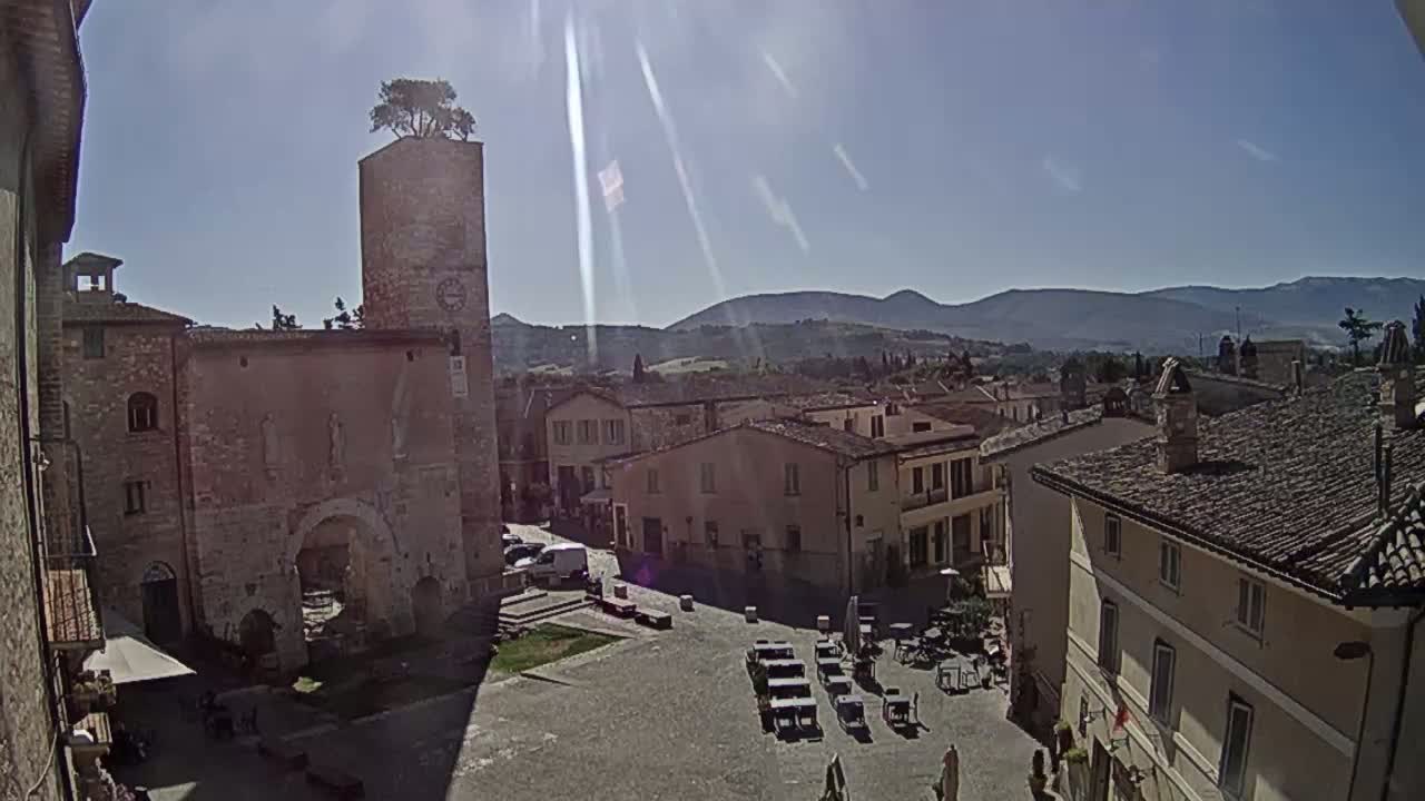 A sunny day reveals a town square in a historic Italian village, surrounded by stone buildings and a clock tower topped with a tree, with distant rolling hills visible under a clear sky.