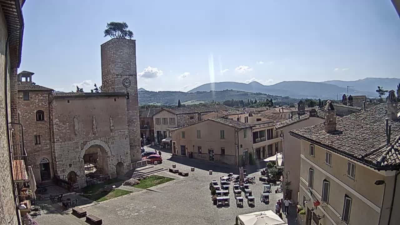 A sunny day reveals a town square in a small Italian town, surrounded by old stone buildings, with a clock tower and distant mountains visible.