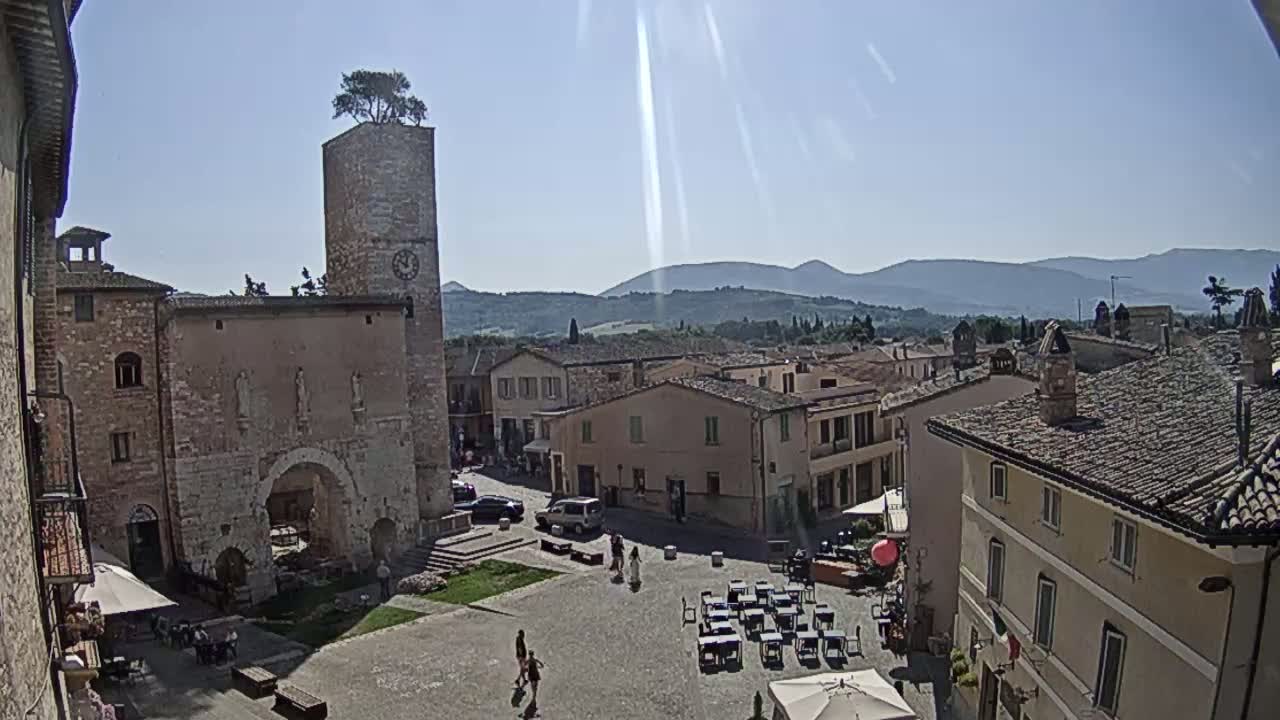 A sunny view of a town square in a small Italian town, featuring stone buildings, a clock tower with a tree on top, and a few people, with mountains visible in the background.