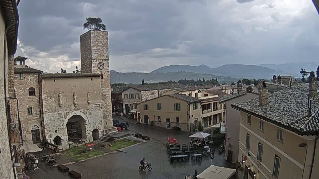 A sunny view of a town square in a small Italian town, featuring stone buildings, a clock tower with a tree on top, and a few people, with mountains visible in the background.