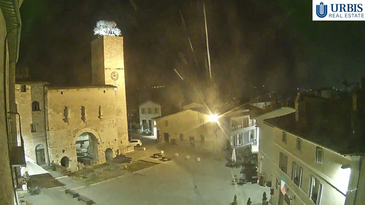 A historic town square is illuminated at night by streetlights and building lights, showcasing a tall stone clock tower topped with a tree and an ancient arched gateway, all under rainy conditions.
