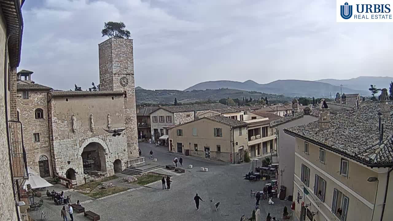 A high-angle view captures a lively historic town square featuring an ancient stone clock tower with a tree on top, surrounded by traditional buildings and distant rolling hills, all under a cloudy sky.