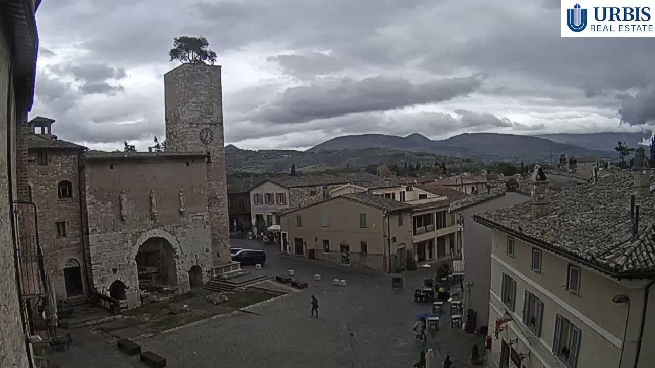 A high-angle view captures a lively historic town square featuring an ancient stone clock tower with a tree on top, surrounded by traditional buildings and distant rolling hills, all under a cloudy sky.