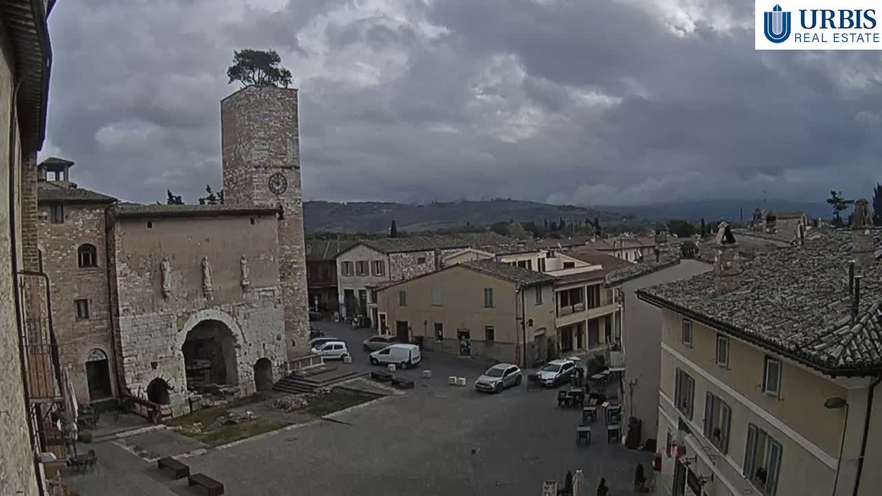 The image displays an ancient town square featuring a prominent stone clock tower with a tree on its summit, historic stone buildings with archways, parked vehicles, and distant rolling hills, all under a heavily overcast and somber sky.