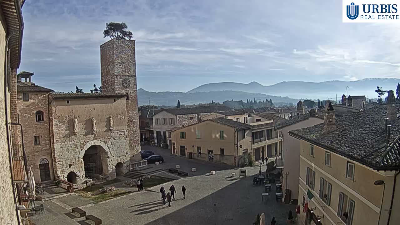 A bustling historic European town square is visible on a bright, partly cloudy day, featuring an ancient stone building with an archway, a tall clock tower topped with a tree, traditional buildings, and distant hazy mountains.