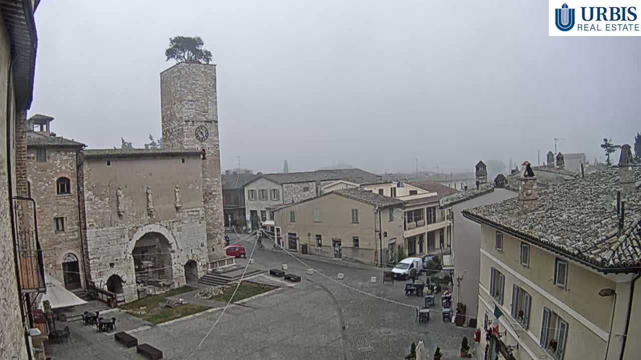 A hazy, overcast sky hangs over an old European town square featuring a stone clock tower with a tree growing from its top, historic buildings, and outdoor cafe tables on the cobbled ground.