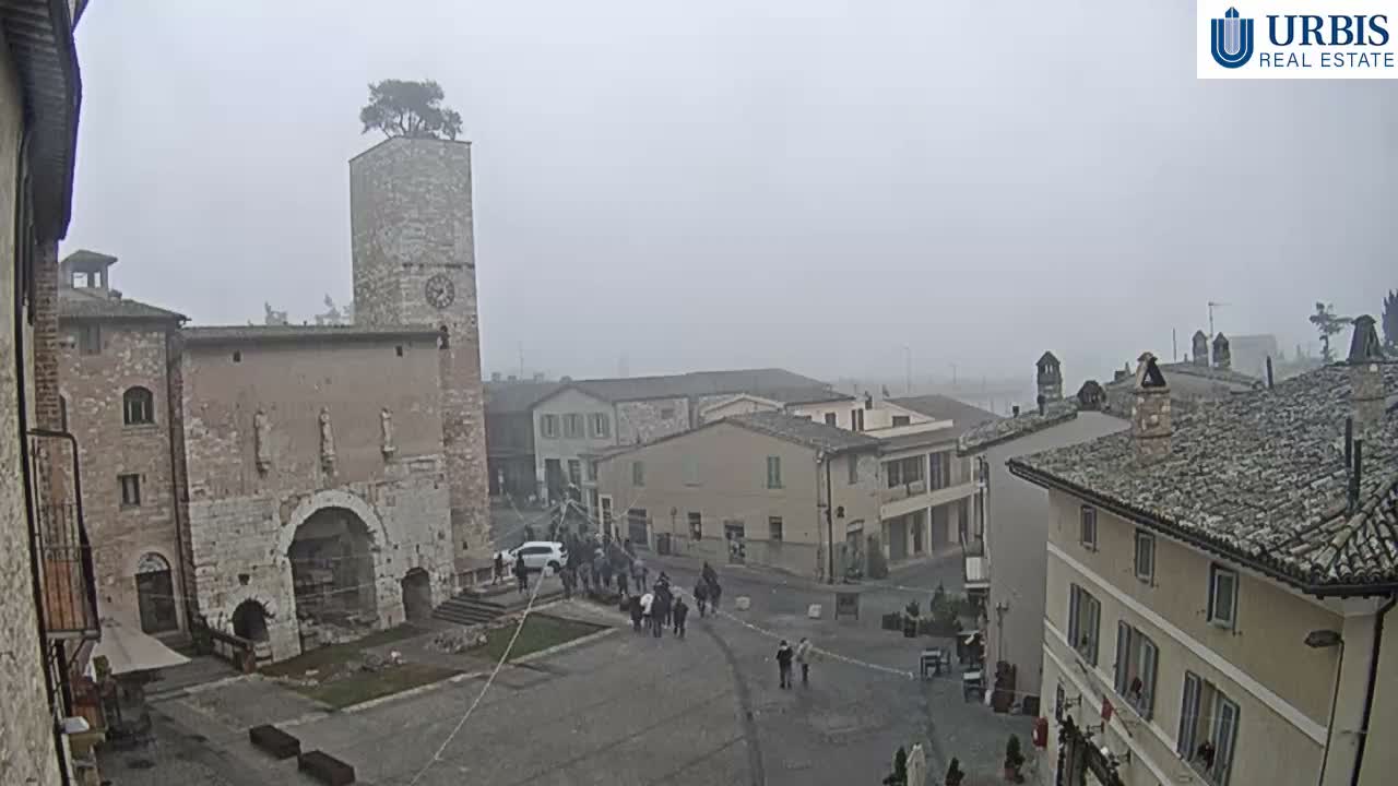 A hazy, overcast sky hangs over an old European town square featuring a stone clock tower with a tree growing from its top, historic buildings, and outdoor cafe tables on the cobbled ground.
