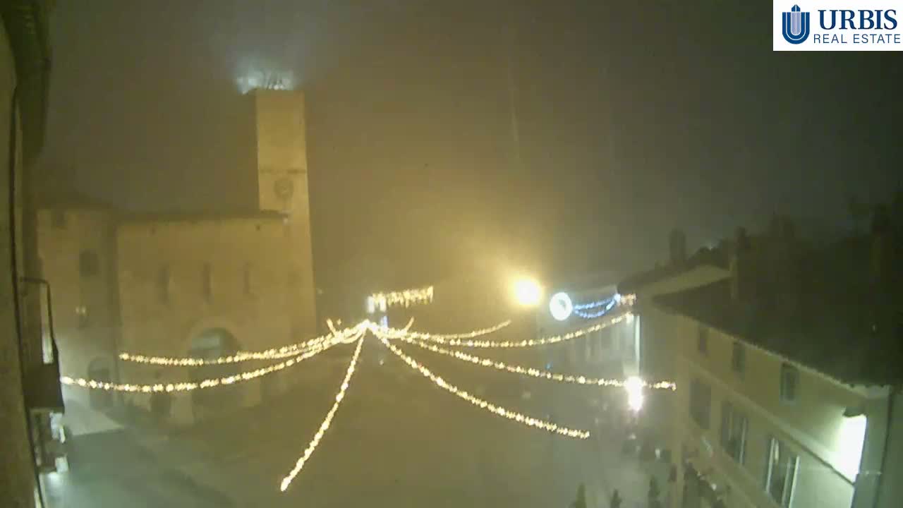 A hazy, overcast sky hangs over an old European town square featuring a stone clock tower with a tree growing from its top, historic buildings, and outdoor cafe tables on the cobbled ground.