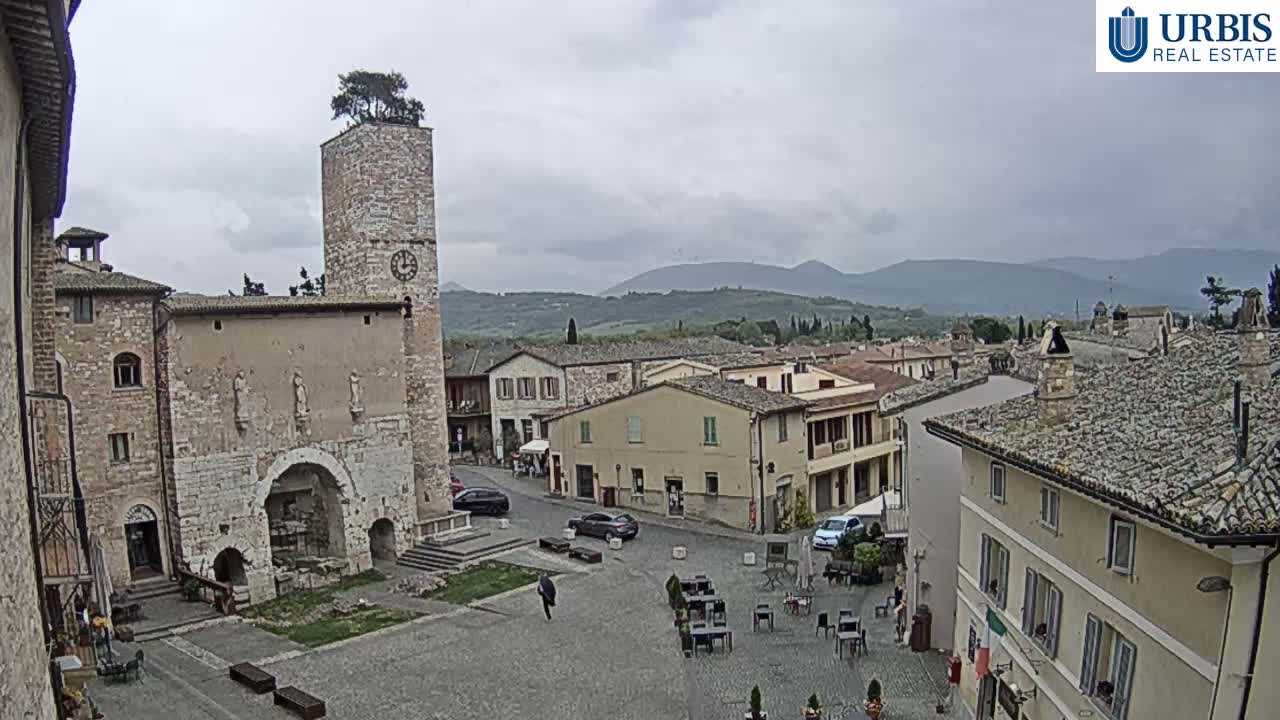 A hazy, overcast sky hangs over an old European town square featuring a stone clock tower with a tree growing from its top, historic buildings, and outdoor cafe tables on the cobbled ground.
