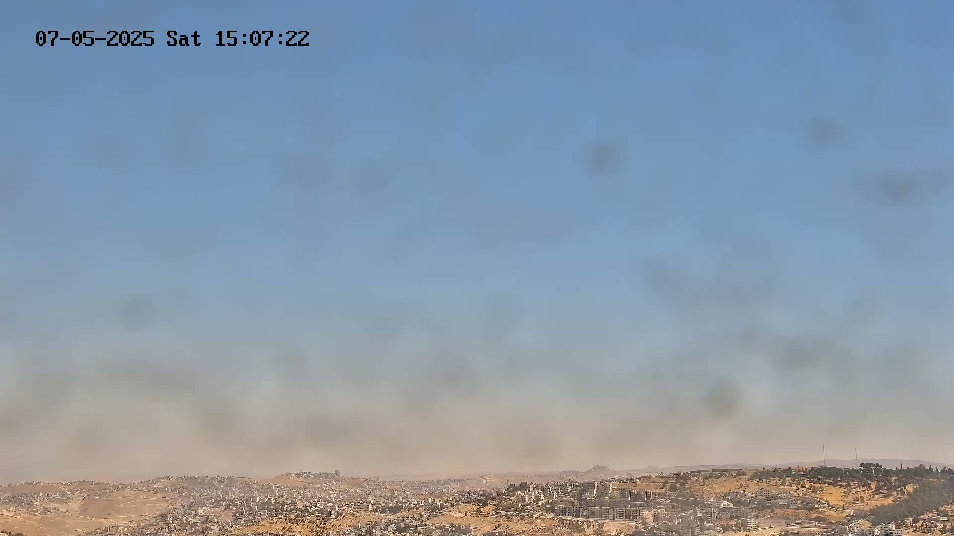A hazy, dusty sky hangs over a sprawling city nestled in a hilly, arid landscape.
