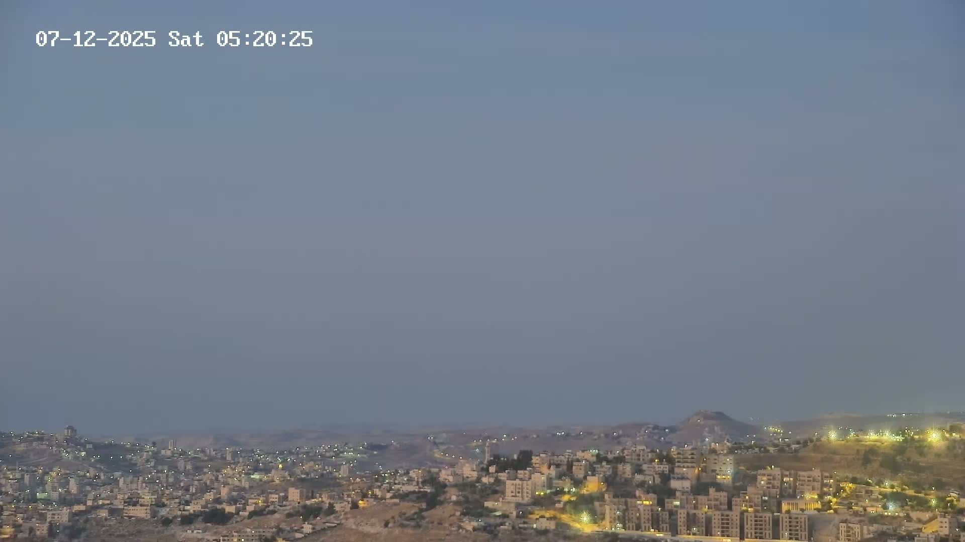 A hazy, dusty sky hangs over a sprawling city nestled in a hilly, arid landscape.