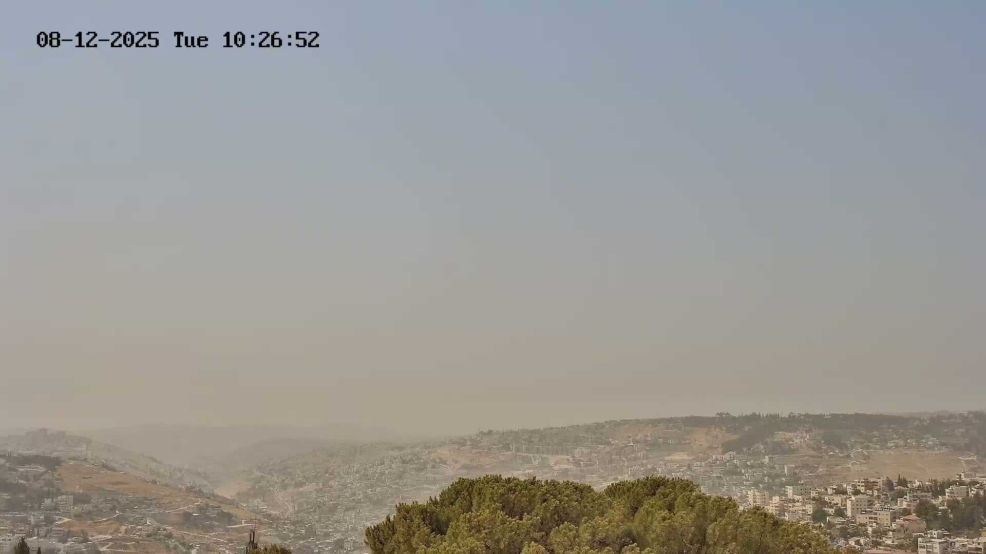 A hazy, dusty view of a sprawling city nestled in a valley, surrounded by hills.