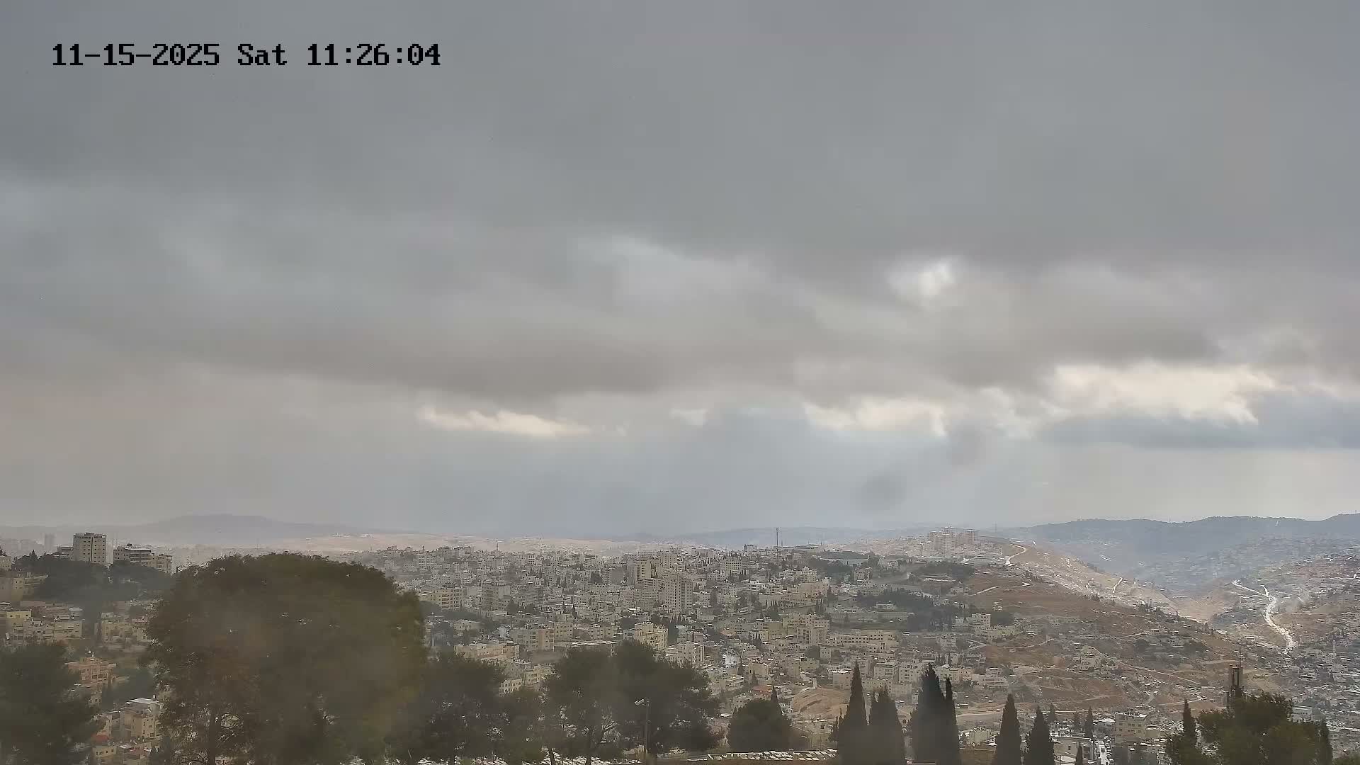 A sprawling city built into rolling hills and valleys is seen under a heavily overcast and gloomy sky.