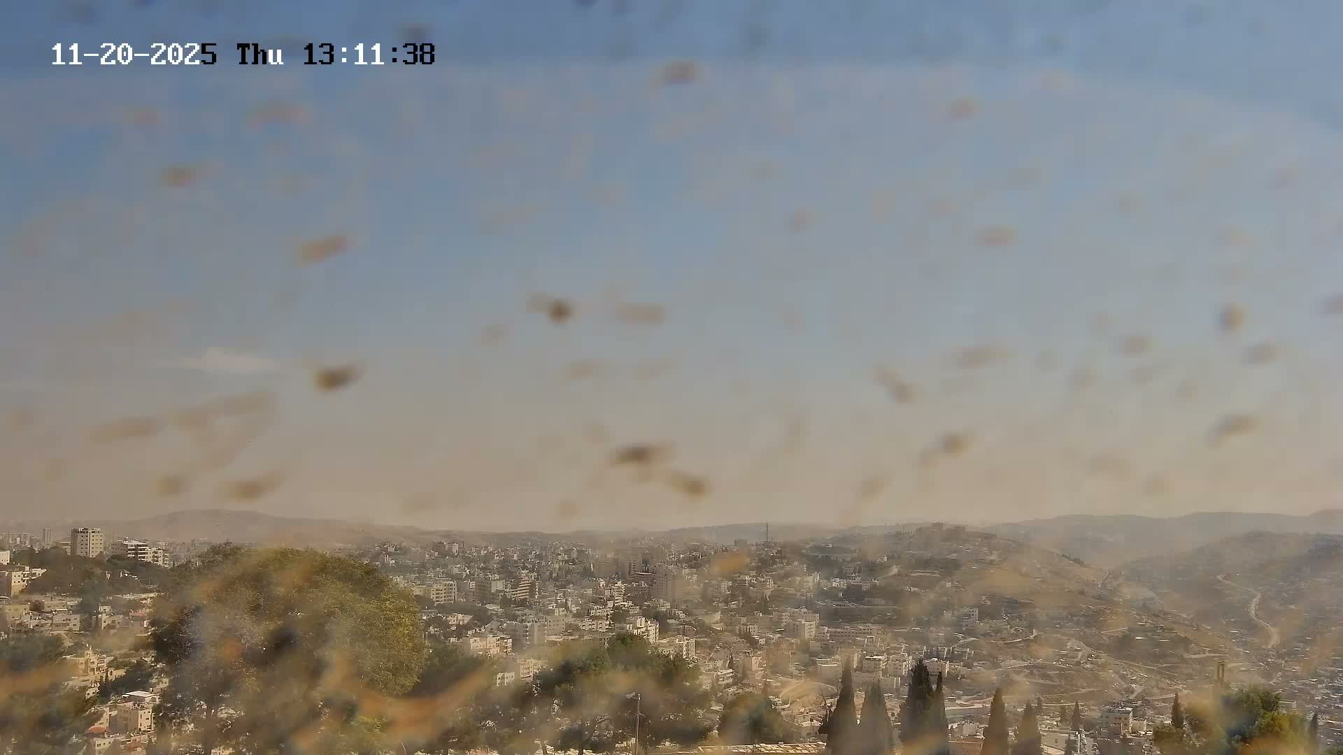 A hazy, panoramic view captures a sprawling city of buildings nestled among hills and distant mountains under a bright but dusty blue sky, significantly obscured by numerous blurry spots on the camera lens.