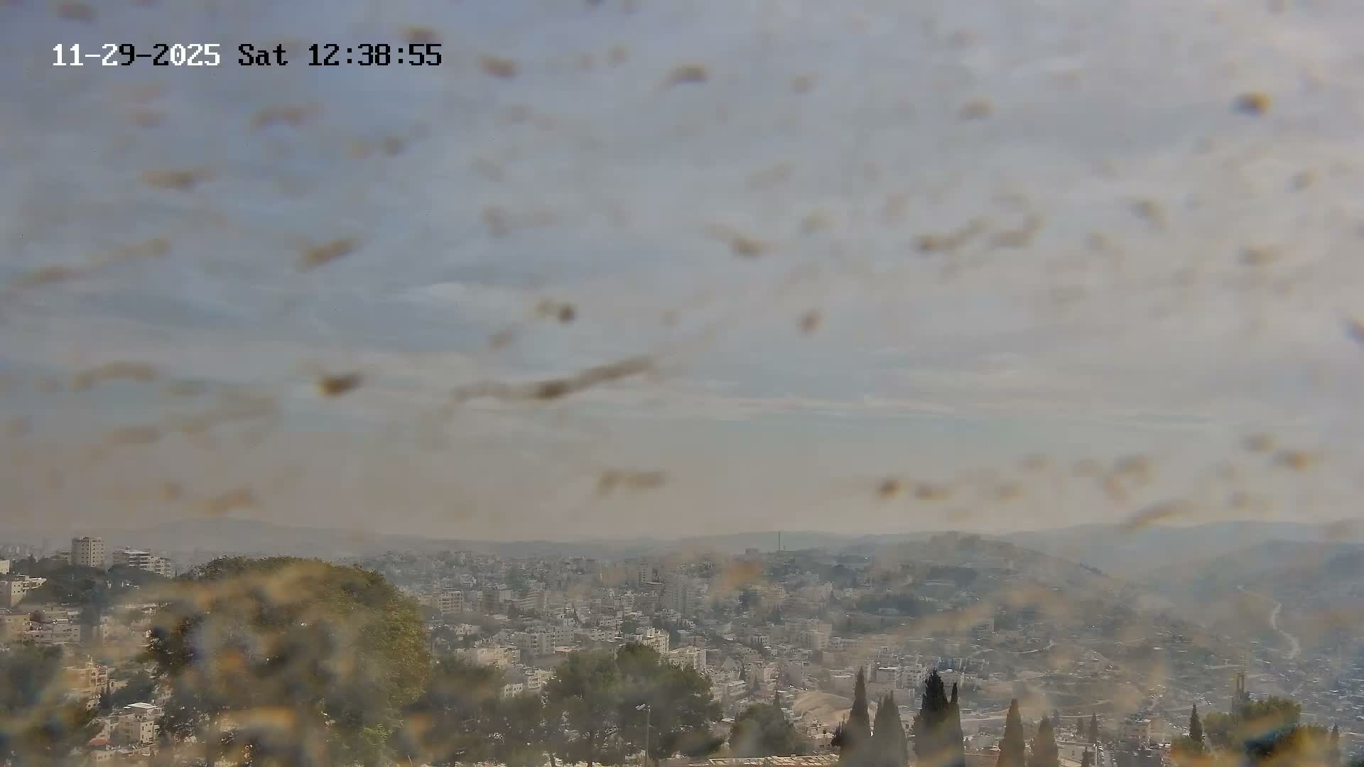 An outdoor scene reveals a sprawling city across rolling hills under a hazy, partly cloudy sky, viewed through a camera lens speckled with numerous debris or water spots.