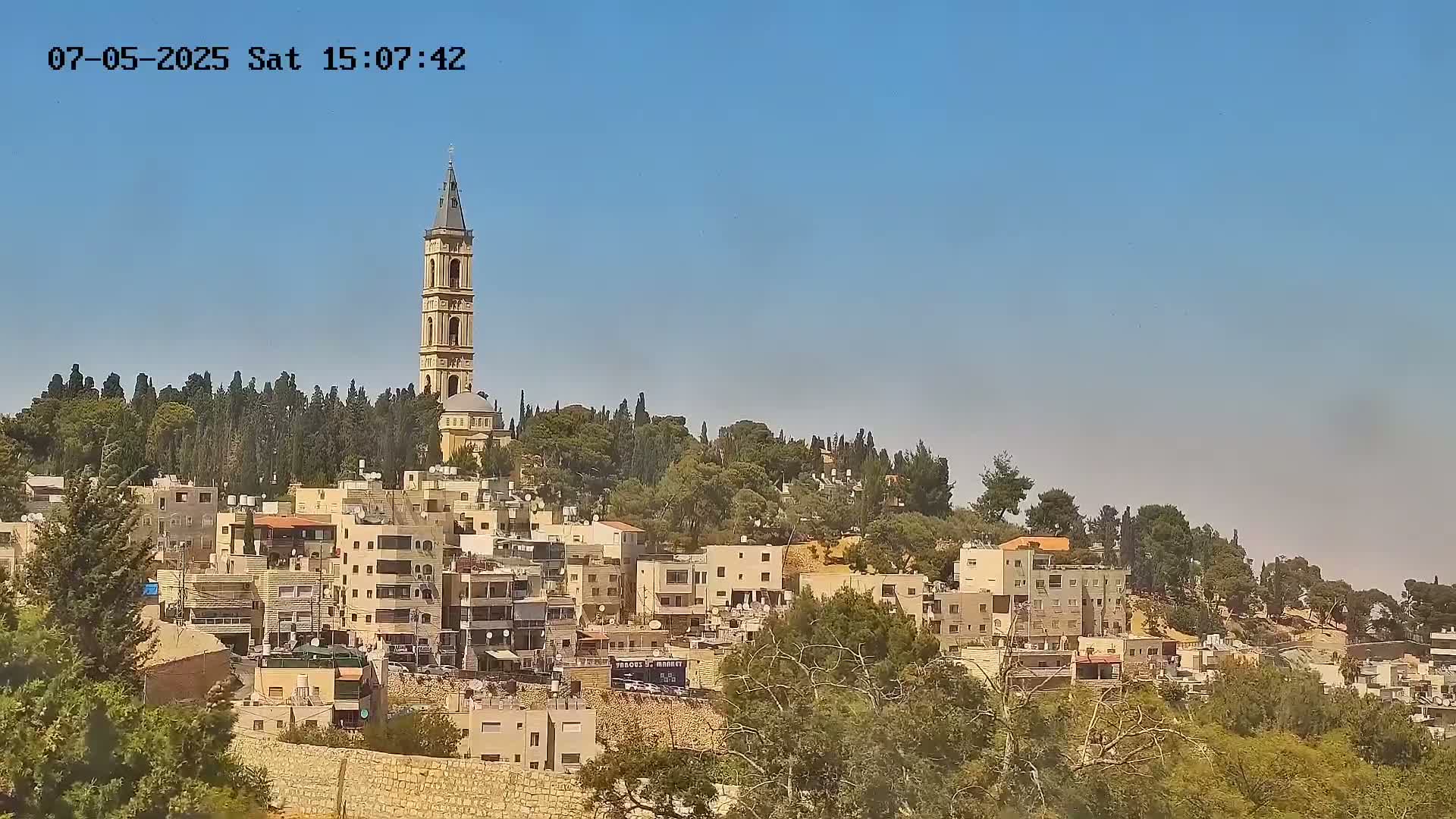 A tall beige tower stands atop a hill overlooking a town of low-rise buildings under a clear blue sky.