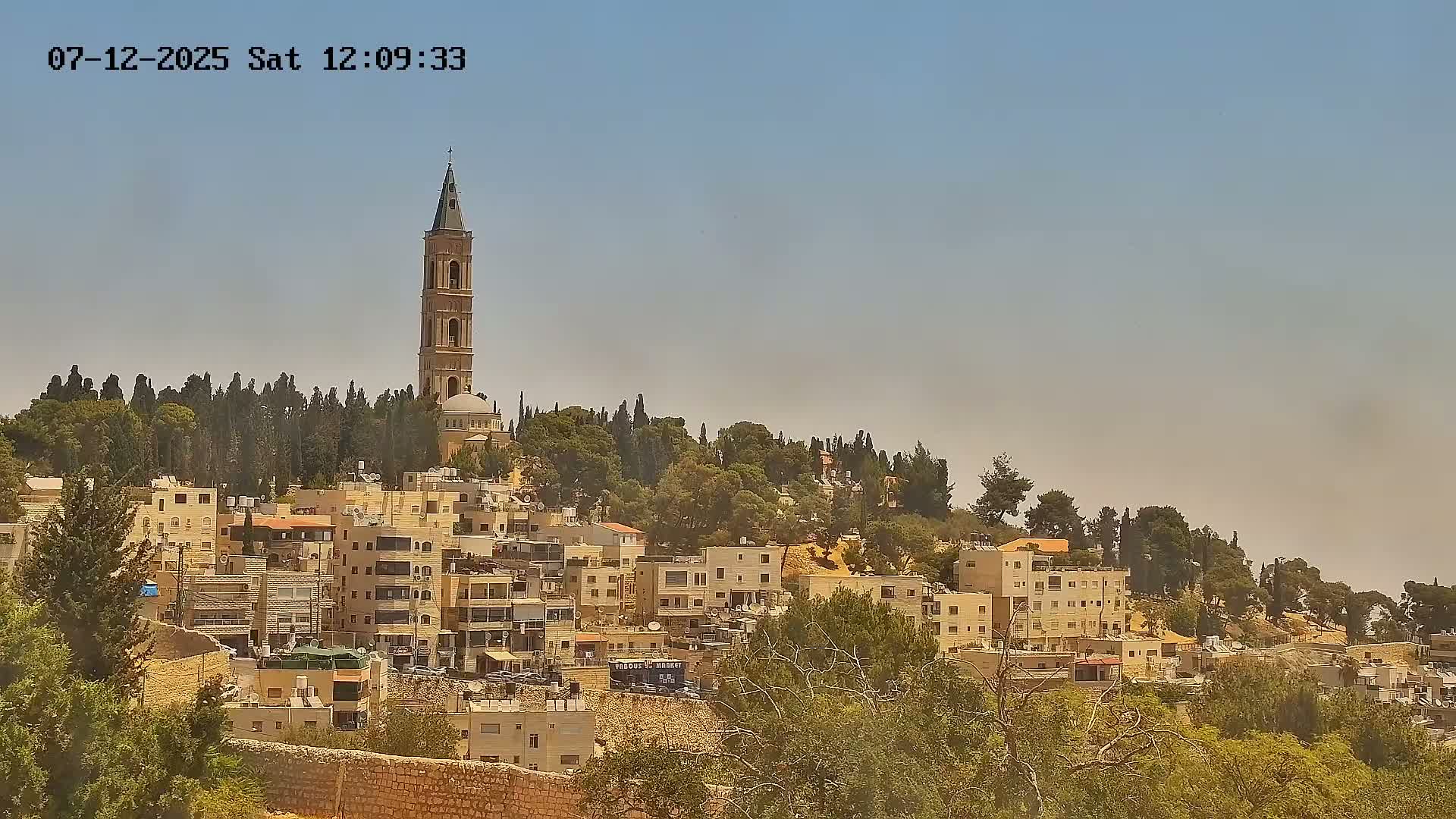A tall beige tower stands atop a hill overlooking a town of low-rise buildings under a clear blue sky.