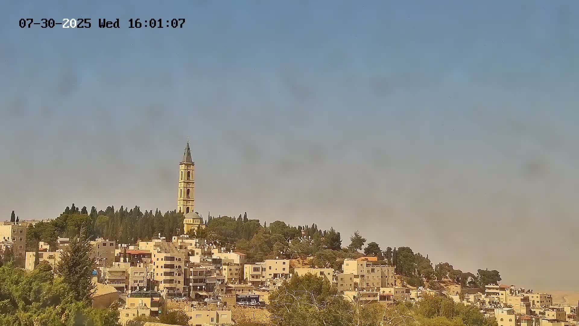 A tall, slender bell tower rises above a hilltop village of light-colored buildings under a clear, hazy sky.