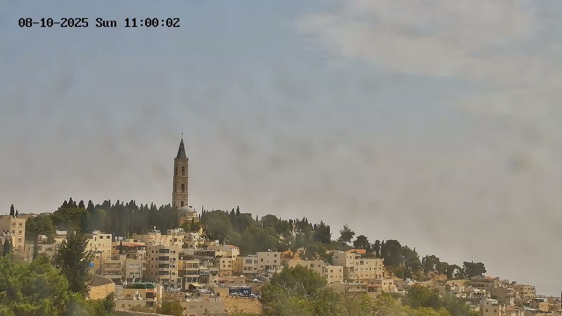 A hazy sky hangs over a hillside town with low buildings, and a tall, slender church tower is visible on the hilltop.