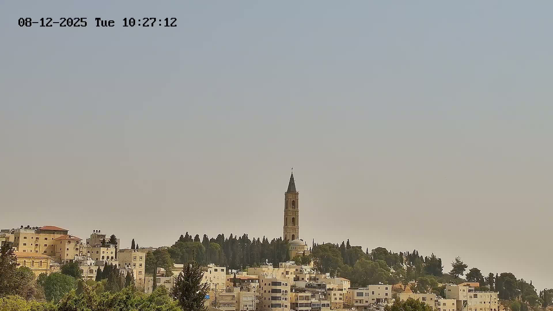 A tall, light-beige tower rises above a hill covered in trees and light-colored buildings under a hazy sky.