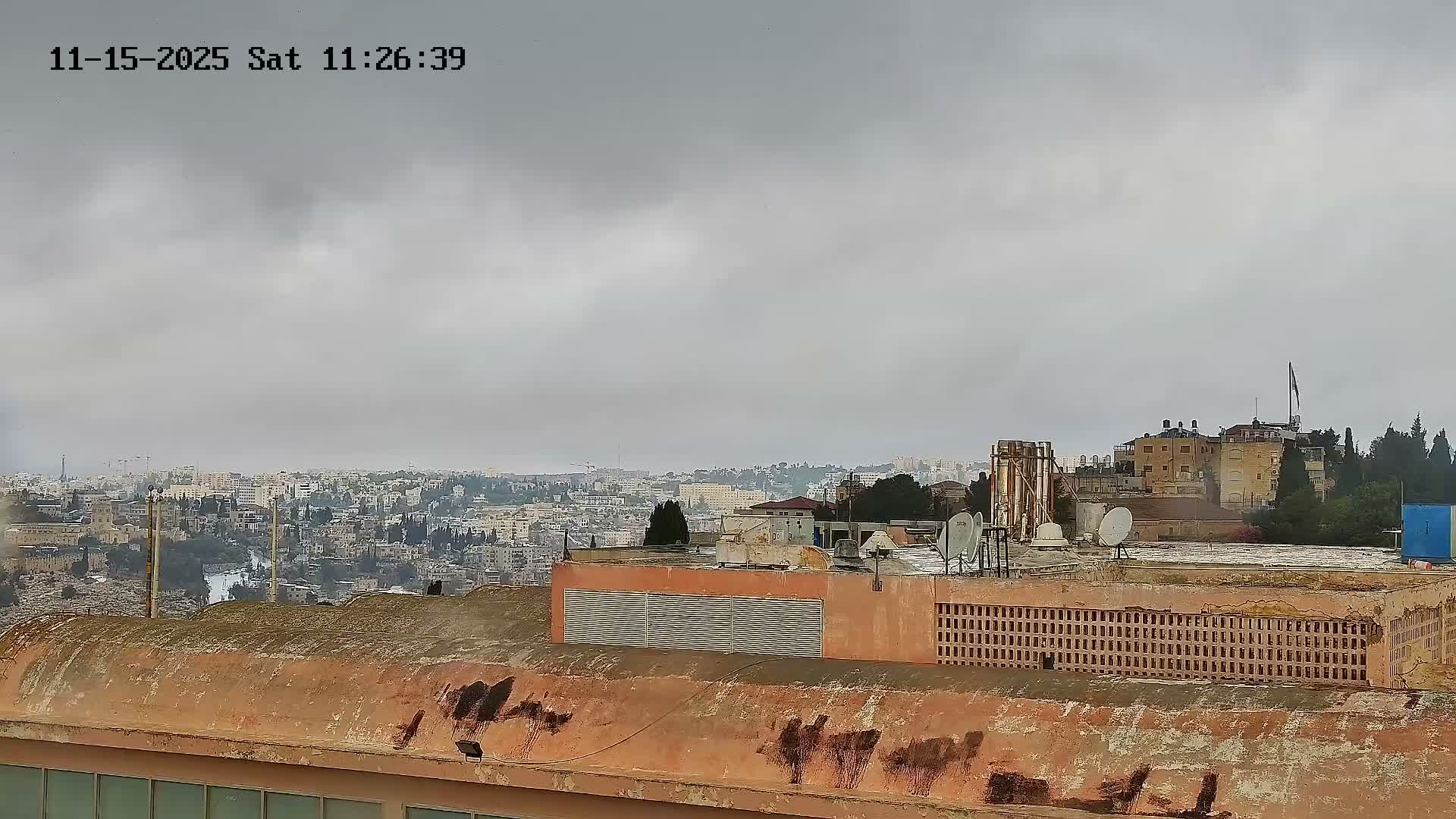 On an overcast day, a sprawling cityscape featuring numerous buildings, some green areas, and a distant body of water is viewed from a weathered orange rooftop covered in patches of dark discoloration, satellite dishes, and vents.