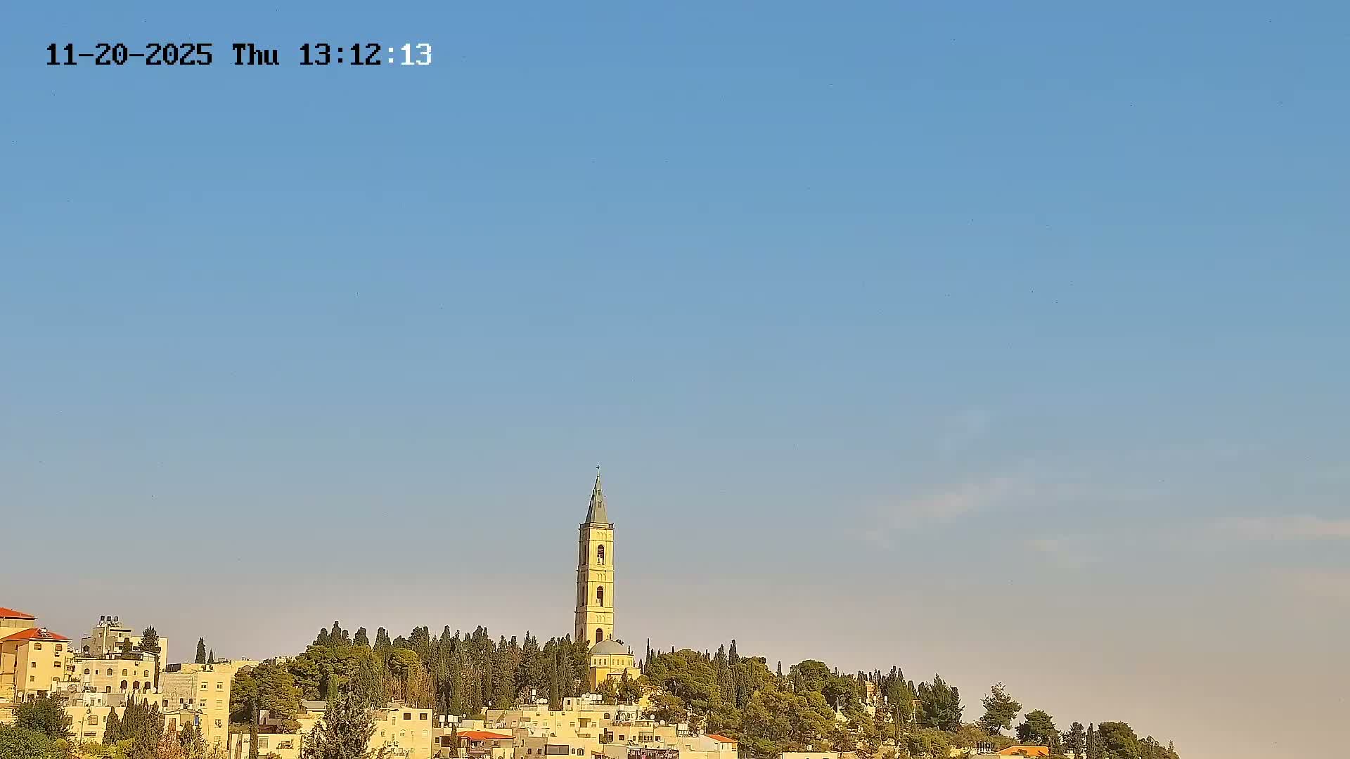 A sunny daytime view shows a tall, light-colored bell tower with a pointed roof rising above a densely wooded hill, flanked by buildings, all set against a clear blue sky.