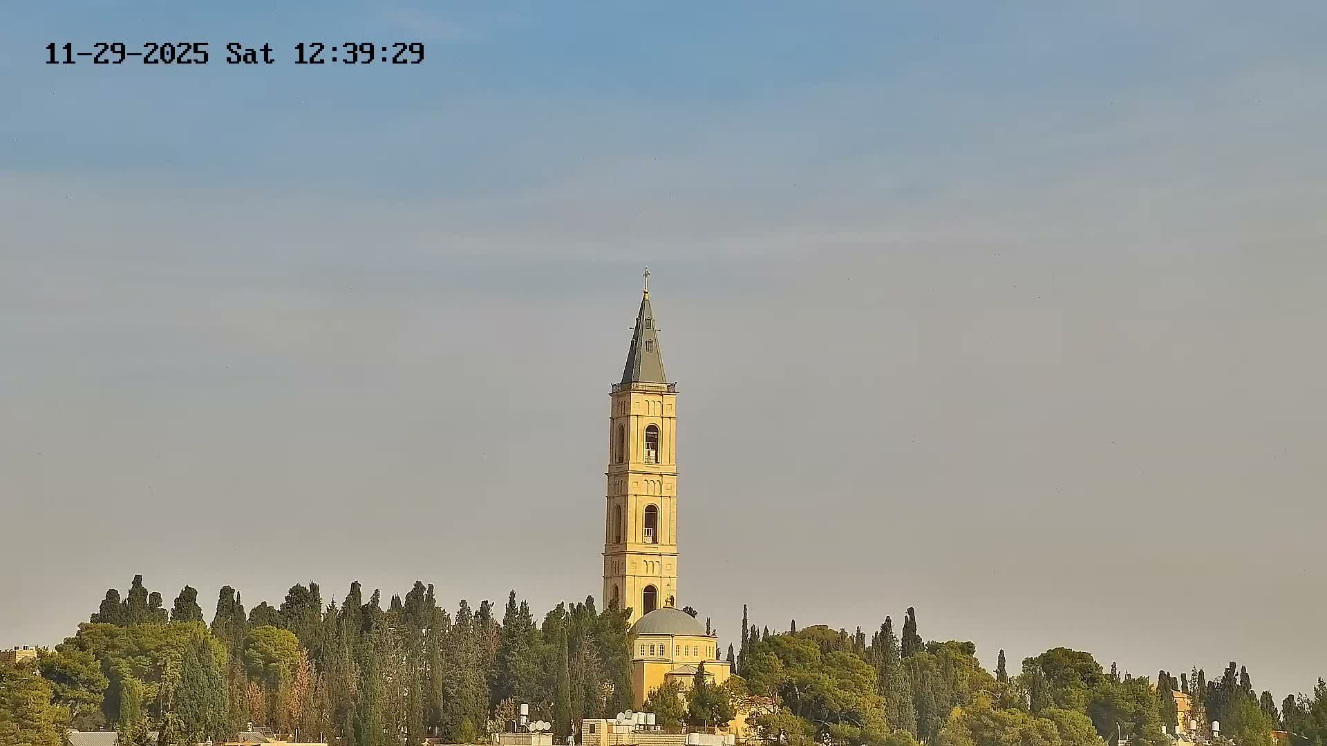 A tall, yellow church tower with a pointed spire and cross rises above a domed building and a dense cluster of green trees, all set against a slightly hazy blue sky.