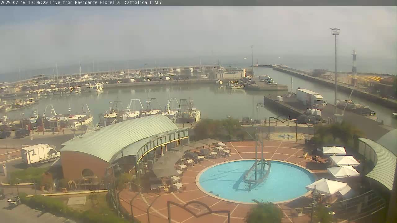 A hazy view of a harbor filled with numerous boats, alongside a plaza containing a circular pool and outdoor seating under umbrellas.