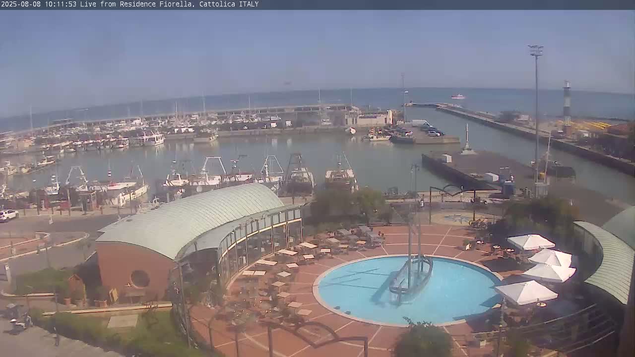 A harbor filled with numerous boats is visible under a clear sky, adjacent to a plaza featuring an outdoor cafe and a circular pool with a ship sculpture.
