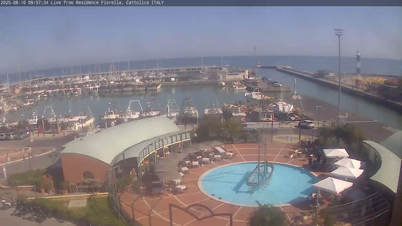 A harbor filled with numerous boats is visible under a sunny sky, overlooking a plaza with a circular fountain and outdoor seating.