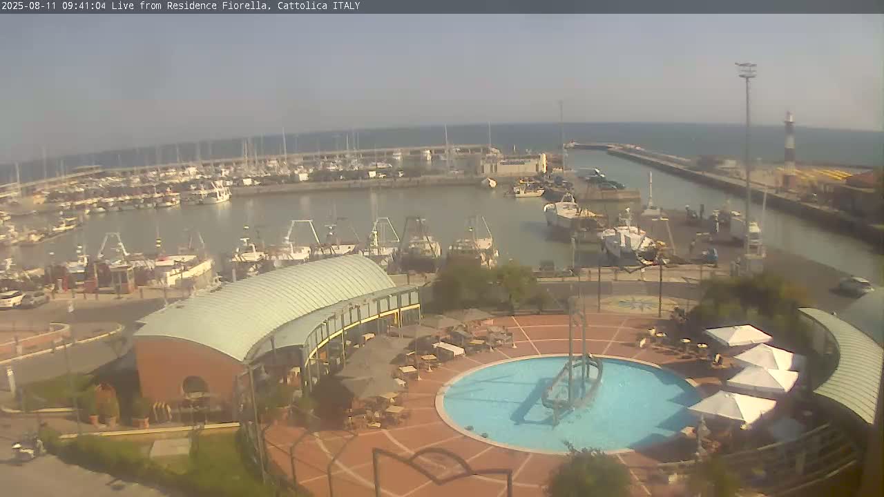 A harbor filled with numerous boats is visible from an elevated vantage point overlooking a patio area with a circular pool and several umbrellas on a sunny day.