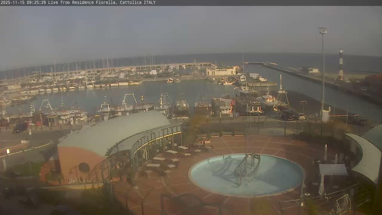 An aerial view captures a bustling harbor filled with numerous boats, from fishing vessels to sailboats, bordered by a modern curved building and an outdoor plaza featuring a circular pool, all under an overcast and hazy sky.