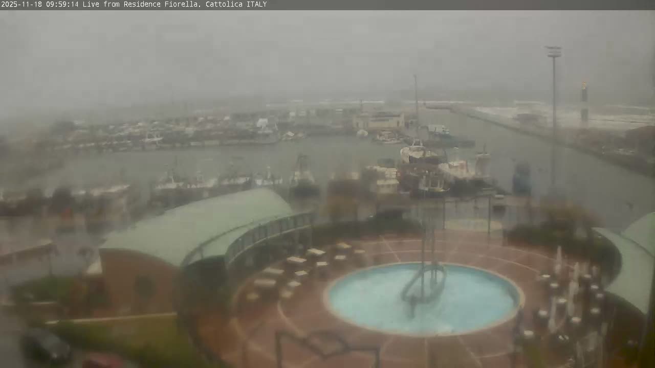 An aerial view captures a bustling harbor filled with numerous boats, from fishing vessels to sailboats, bordered by a modern curved building and an outdoor plaza featuring a circular pool, all under an overcast and hazy sky.