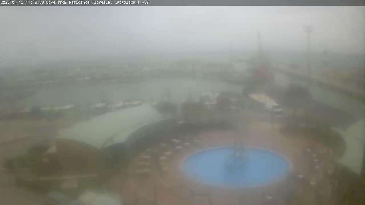 A hazy and overcast view reveals a harbor teeming with numerous boats along a wide waterway, bordered by a circular plaza featuring a boat sculpture fountain and modern buildings in the foreground.