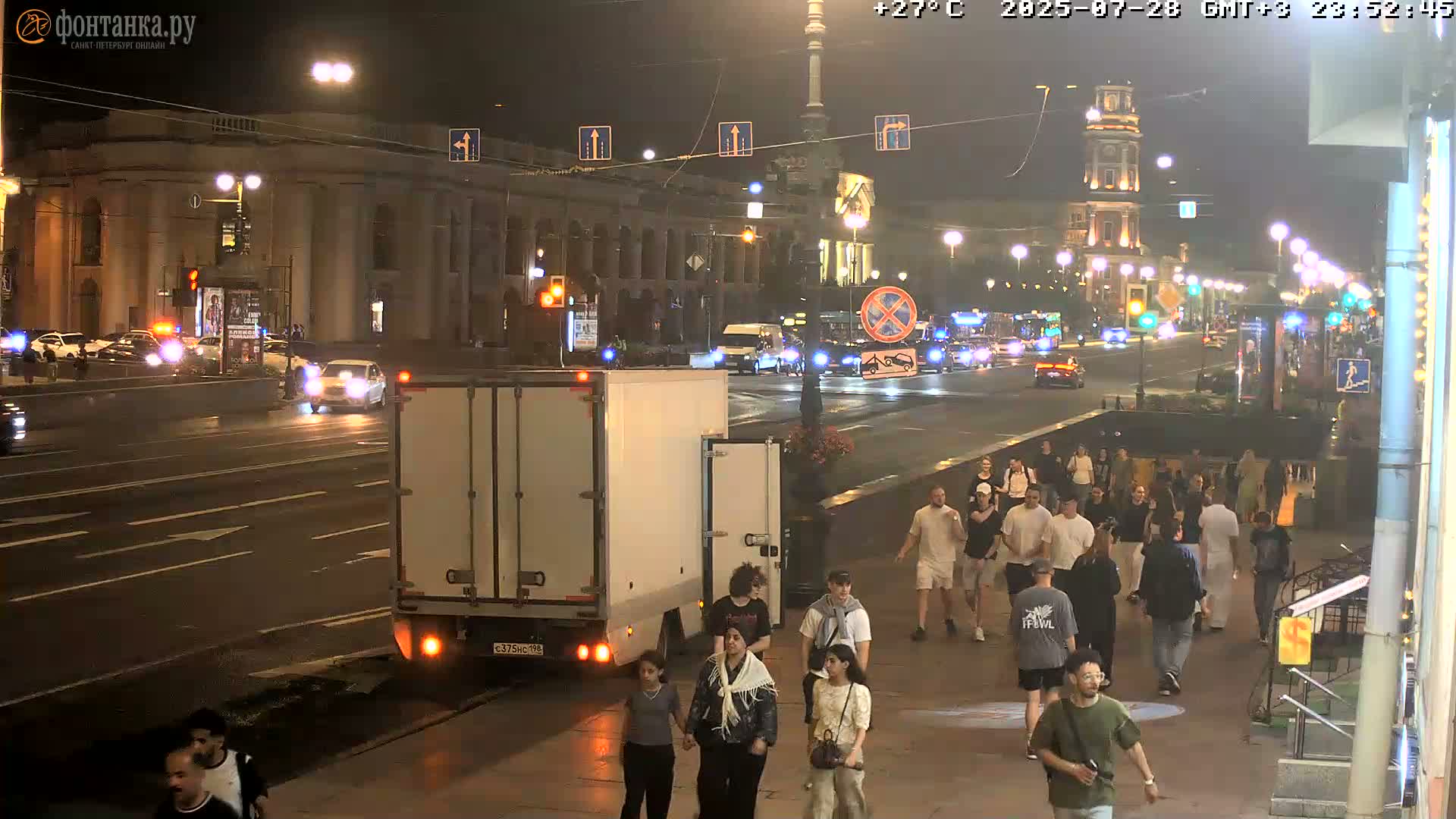 A nighttime city street scene shows a group of pedestrians walking past a parked truck, with cars driving on a road lined with buildings under streetlights.