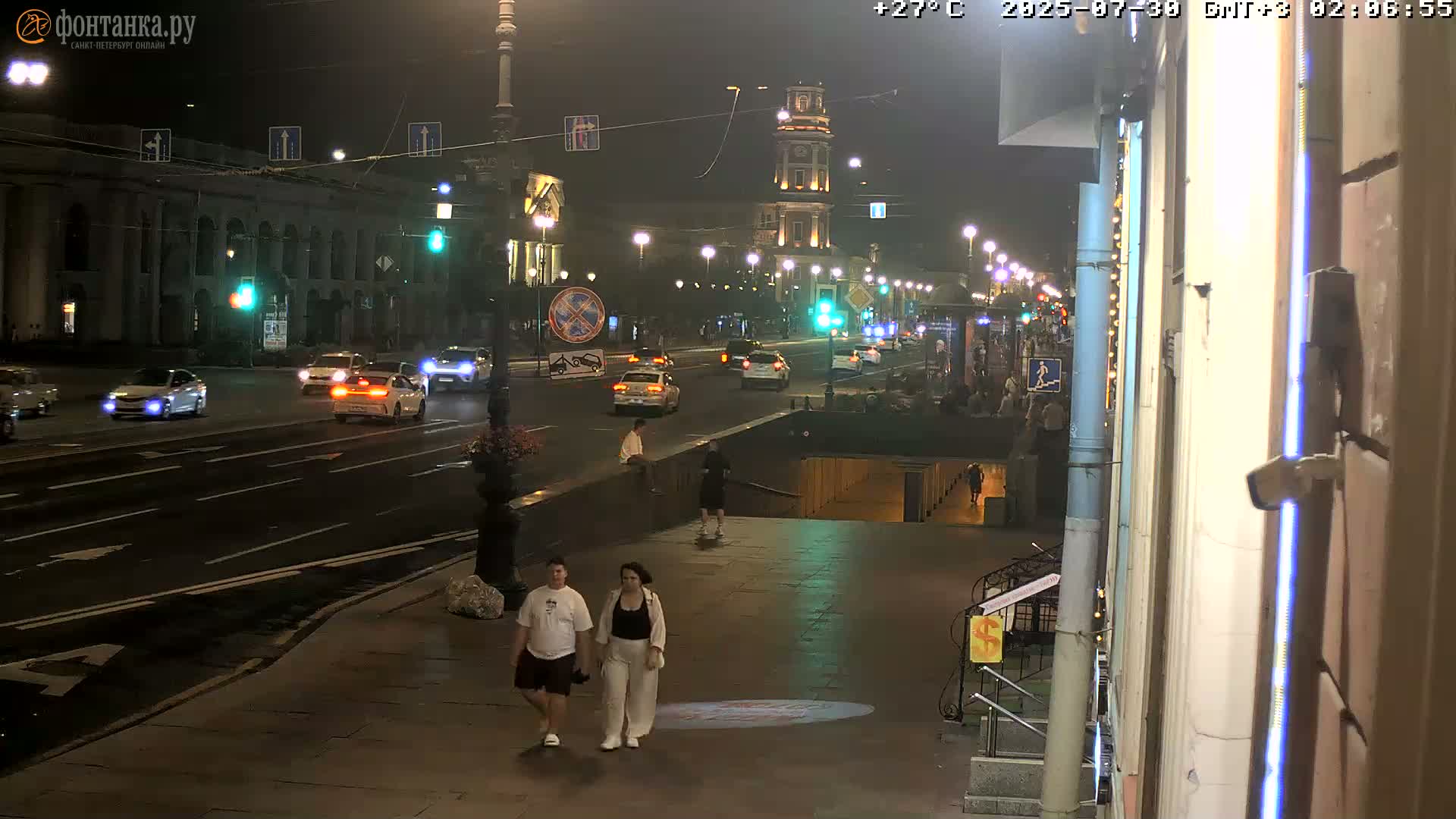 A nighttime city street scene shows a group of pedestrians walking past a parked truck, with cars driving on a road lined with buildings under streetlights.