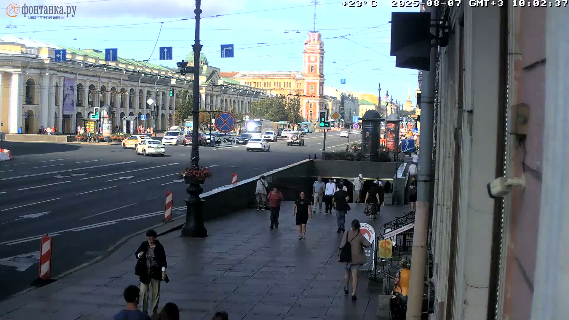 Pedestrians walk on a city sidewalk near a street with cars and buildings under a sunny sky.
