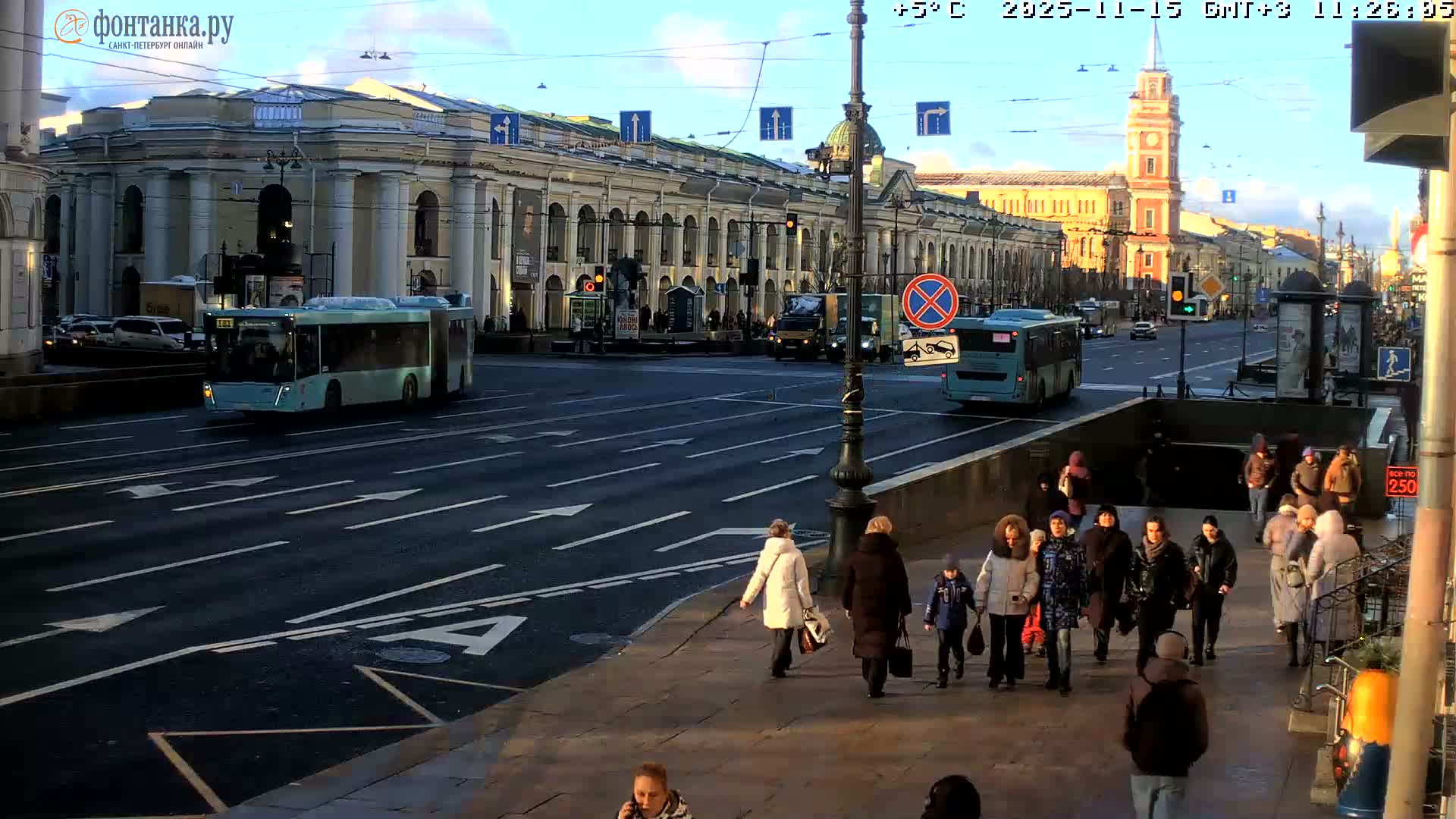 A bustling city street bathed in bright sunlight features multiple lanes of traffic with buses and cars, numerous pedestrians in winter attire on the sidewalk and entering a subway station, all set against a backdrop of grand classical buildings and a distant red and white clock tower under a clear blue sky.