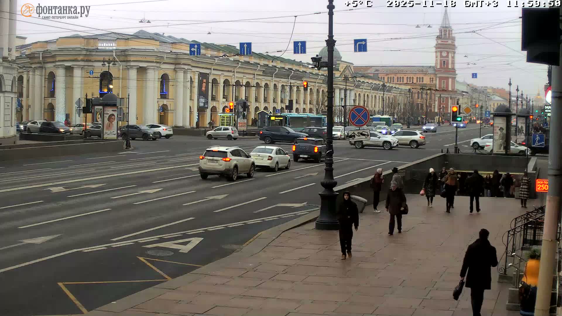 A bustling city street bathed in bright sunlight features multiple lanes of traffic with buses and cars, numerous pedestrians in winter attire on the sidewalk and entering a subway station, all set against a backdrop of grand classical buildings and a distant red and white clock tower under a clear blue sky.