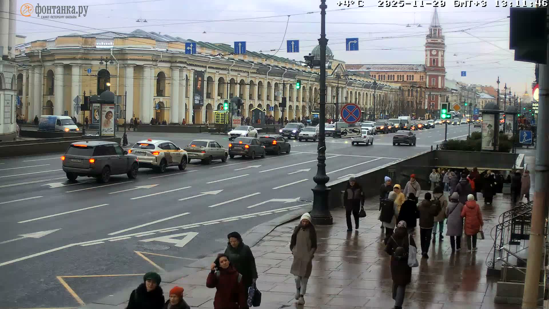 On a cold, overcast day, a wide city street appears wet from recent precipitation, bustling with vehicle traffic and pedestrians bundled in winter clothing along the sidewalks and near grand architectural buildings, with a prominent clock tower in the distance.