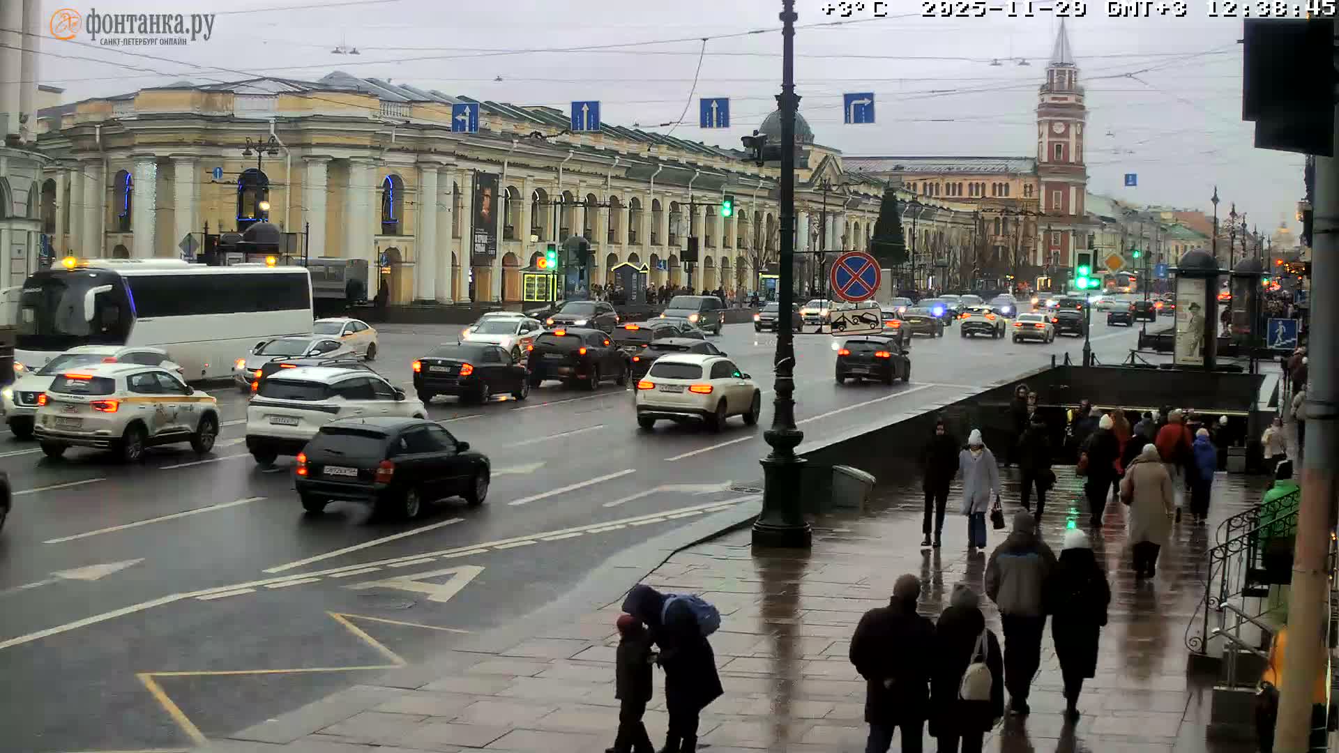 A busy city street is depicted on an overcast and wet day, filled with cars on a glistening road and pedestrians navigating sidewalks and an underground entrance, all against a backdrop of stately classical buildings and a distant clock tower.