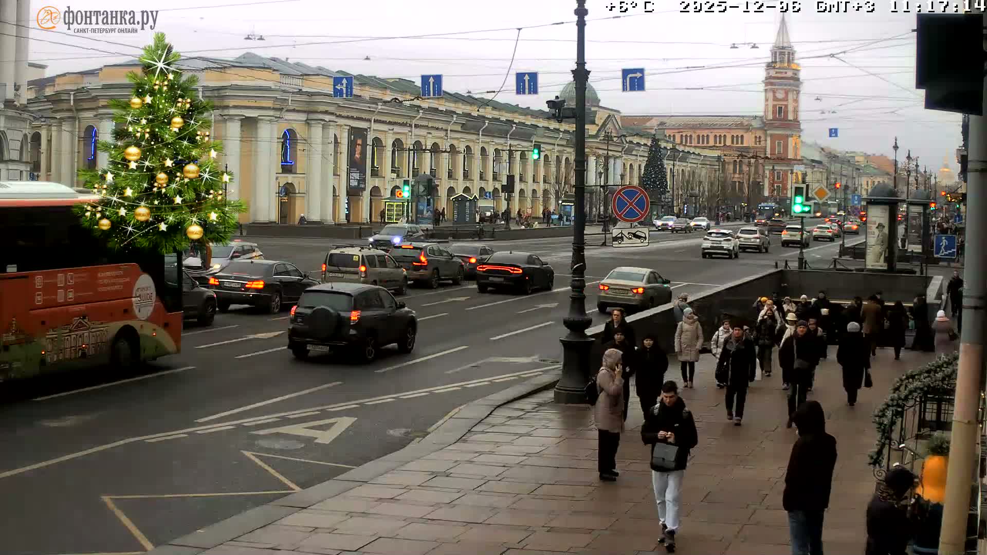 An overcast daytime city scene features a large decorated Christmas tree next to a busy road with numerous cars, a prominent sidewalk filled with pedestrians, and classical buildings with a distant clock tower under a grey sky.