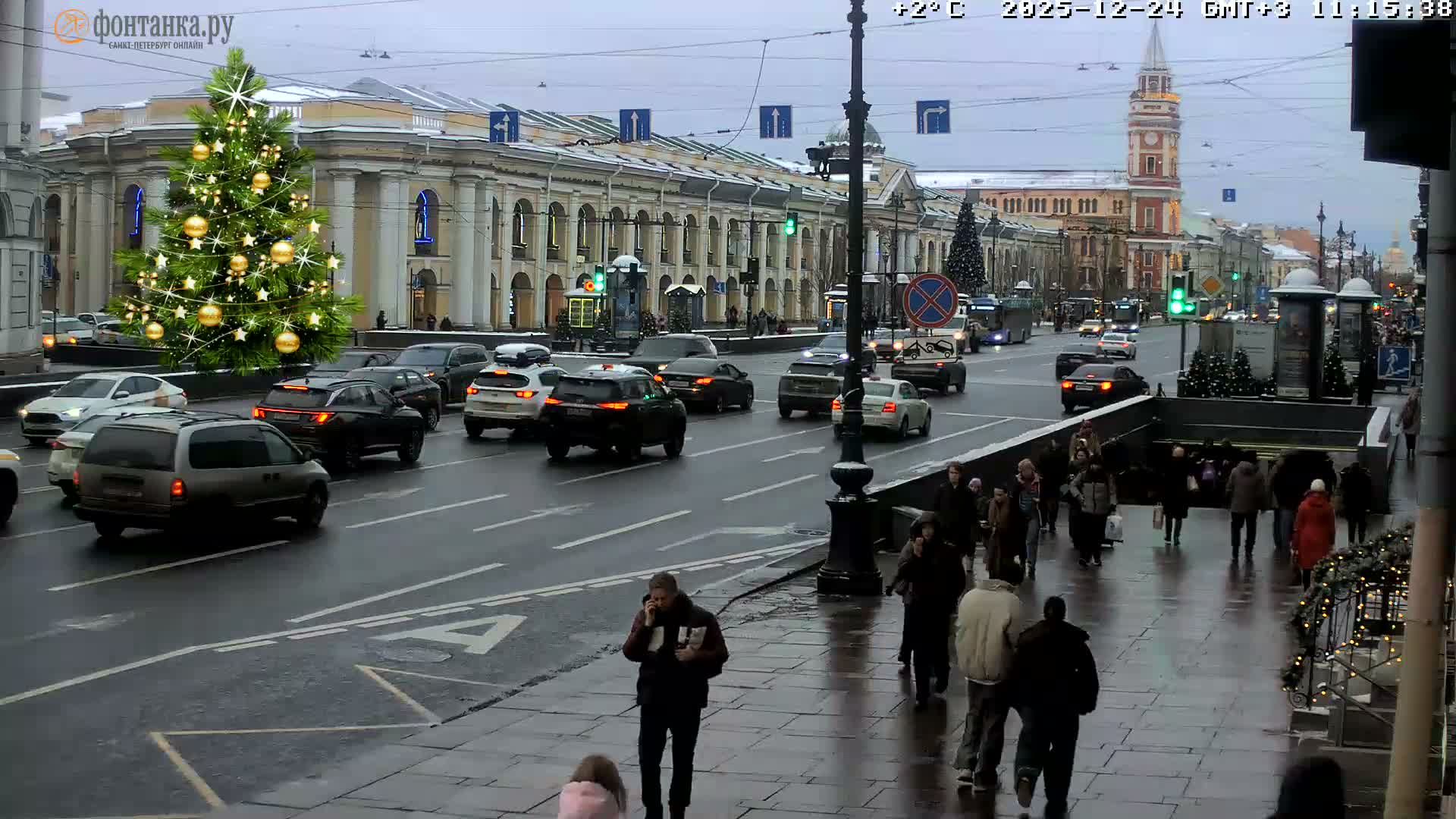 A busy city street is shown on an overcast day with wet roads, featuring a large, decorated Christmas tree on the left, historical buildings lining the thoroughfare, numerous cars moving and parked, and many pedestrians walking on the sidewalks and entering a subway station.