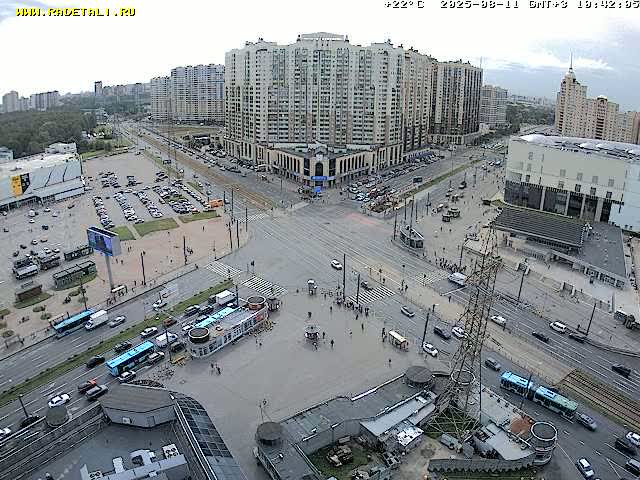 A high-angle, wide view shows a city square with traffic, pedestrians, and several large buildings under partly cloudy skies.